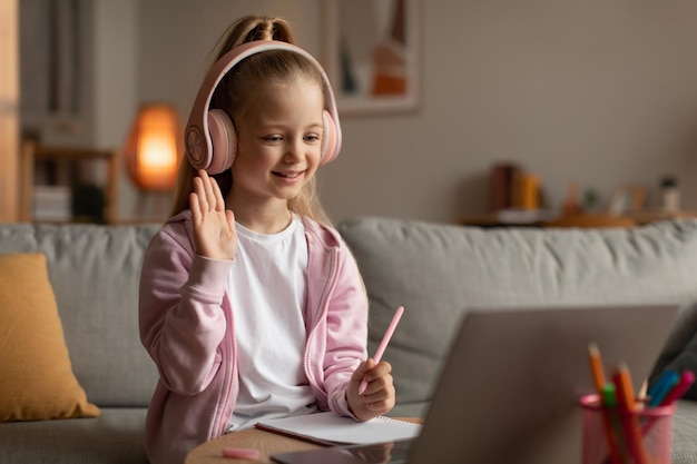 Klein schoolmeisje dat hallo zwaait naar laptop die thuis online leert