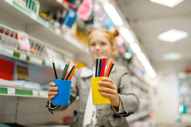 Klein schoolmeisje houdt een bril met potloden vast, winkelt in een kantoorboekhandel