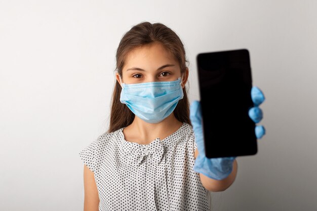 Klein schoolmeisje in medisch masker en handschoenen die mobiele telefoon tonen aan de camera