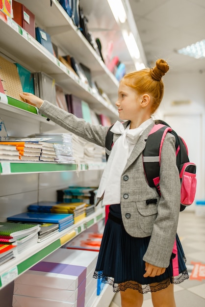 Klein schoolmeisje met rugzak op de plank in de kantoorboekhandel