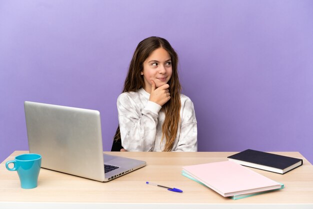 Klein studentenmeisje op een werkplek met een laptop geïsoleerd op een paarse achtergrond die een idee denkt terwijl ze omhoog kijkt