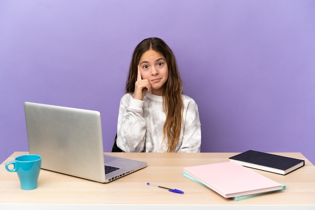 Klein studentenmeisje op een werkplek met een laptop geïsoleerd op een paarse achtergrond die een idee denkt