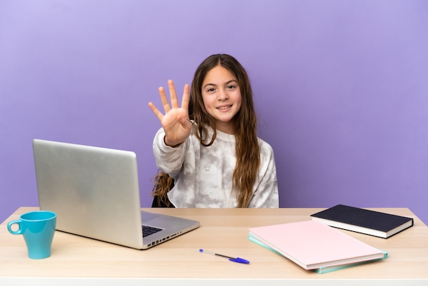 Klein studentenmeisje op een werkplek met een laptop geïsoleerd op een paarse achtergrond gelukkig en vier tellen met vingers