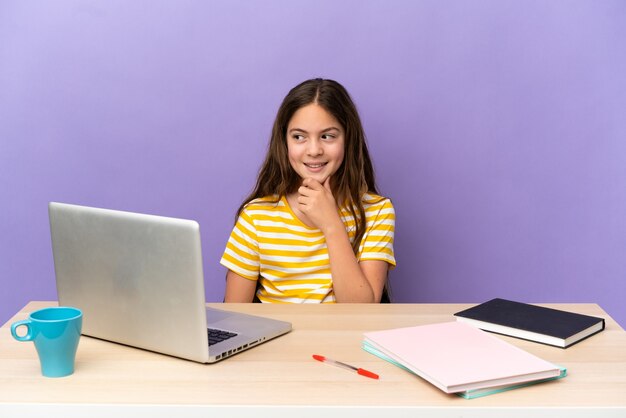 Klein studentenmeisje op een werkplek met een laptop geïsoleerd op een paarse achtergrond, opzij kijkend en glimlachend