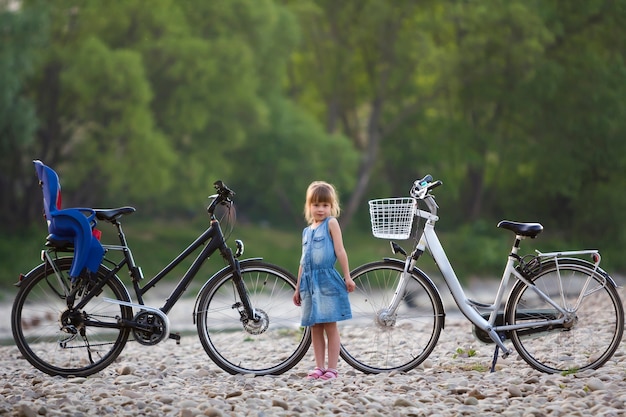 Foto klein vrij blond meisje in blauwe kleding die zich voor witte fiets met emmer en zwarte met kinderzitje op vage groene bomenachtergrond bevinden. actief levensstijl en familierecreatieconcept.