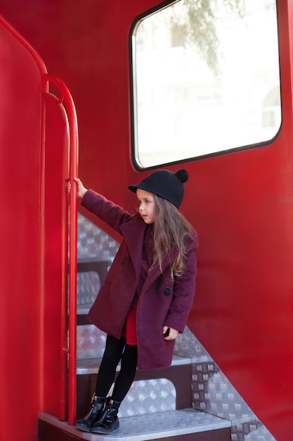 Klein vrolijk meisje in de buurt van de rode engelse bus in een mooie jas en een hoed. reis van het kind. schoolbus. londen rode bus. voorjaar. met de internationale vrouwendag. sinds 8 maart!