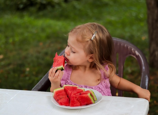 klein vrolijk meisje in paarse kleren eet watermeloen met eetlust, genietend van de zoete smaak