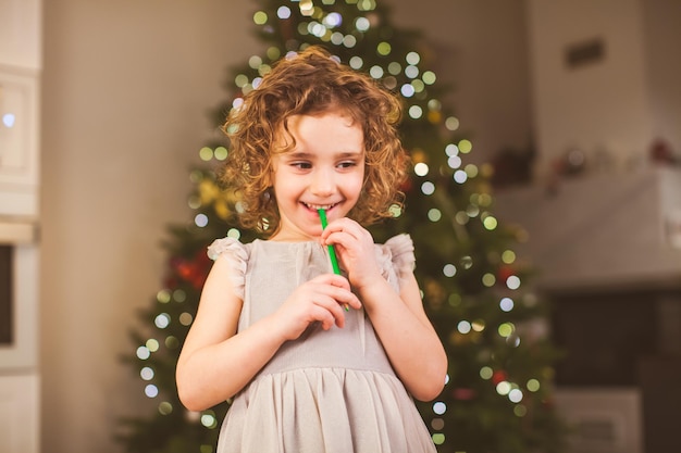 Klein vrolijk meisje met krullend haar staat in de woonkamer met versierde kerstboom op de achtergrond dromen over geschenken met groen potlood in haar mond Kerstconcept Nieuwjaar