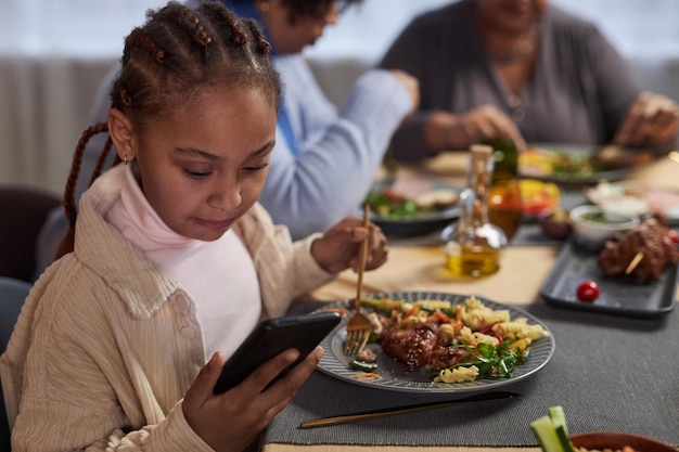 Klein zwart meisje met telefoon aan de eettafel.