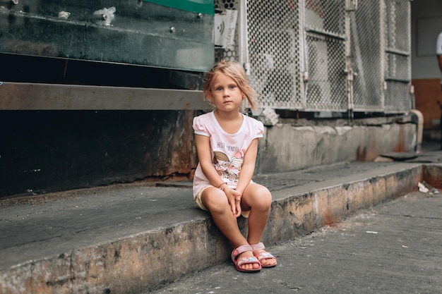 Kleine aantrekkelijke blonde jongen zit op betonnen grond en wacht op iets, portret