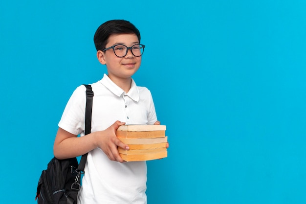 kleine aziatische schooljongen in glazen met rugzak die boeken vasthoudt en glimlacht op blauwe achtergrond