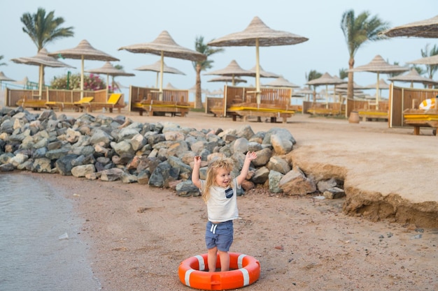 Kleine babyjongen of schattig kind met blij gezicht en blond haar in shirt en korte broek blootsvoets spelen met rode veiligheidsgordel of reddingsgordel op zandstrand met stenen parasols op natuurlijke achtergrond