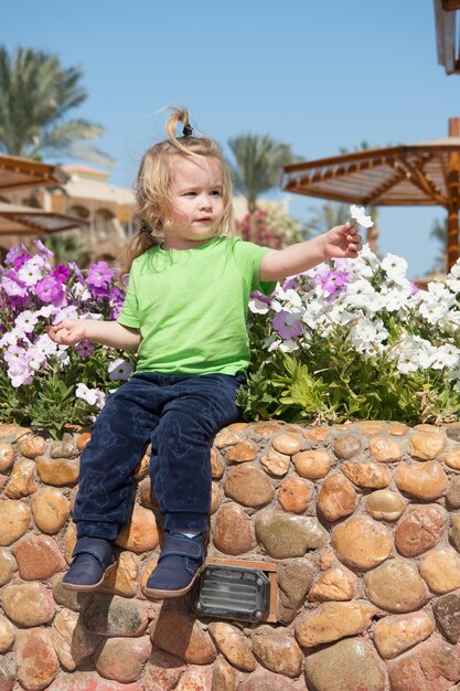 Kleine babyjongen of schattig kind met schattig gezicht en blond haar in groen shirt en broek zittend op steenachtige borstwering spelen met bloemen zonnig buiten op natuurlijke blauwe hemelachtergrond