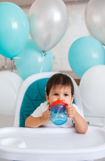 Kleine babyjongen zittend in blauwe kinderstoel thuis op witte keuken en drinkwater uit sippy cup op achtergrond met ballonnen.