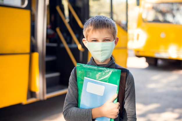 Kleine basisschoolleerling die voor de oranje schoolbus staat en een gezichtsmasker draagt