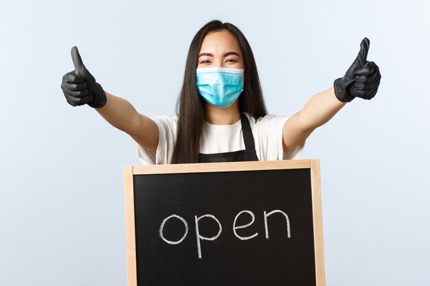 Kleine bedrijven, covid-19 pandemie, preventie van virus en werknemersconcept. Vrolijke Aziatische vrouwelijke winkelmedewerker, cafépersoneel met medisch masker nodigt bezoekers uit, duim omhoog en we zijn open teken