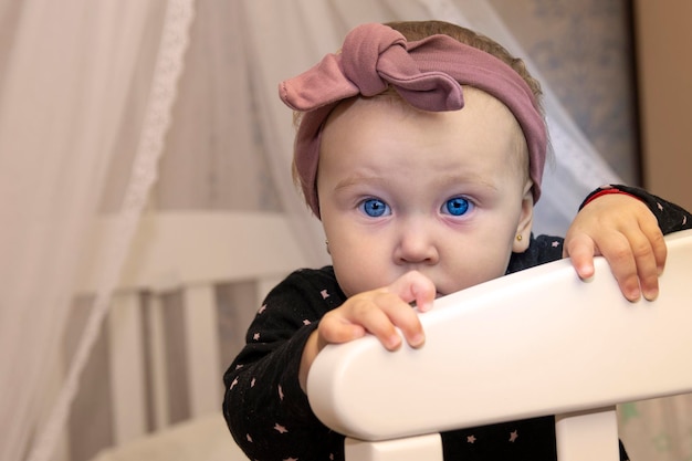 kleine blanke babymeisje gluurt uit de witte wieg. babymeisje 10 maanden oud met mooie blauwe ogen en een rood hoofdbandportret
