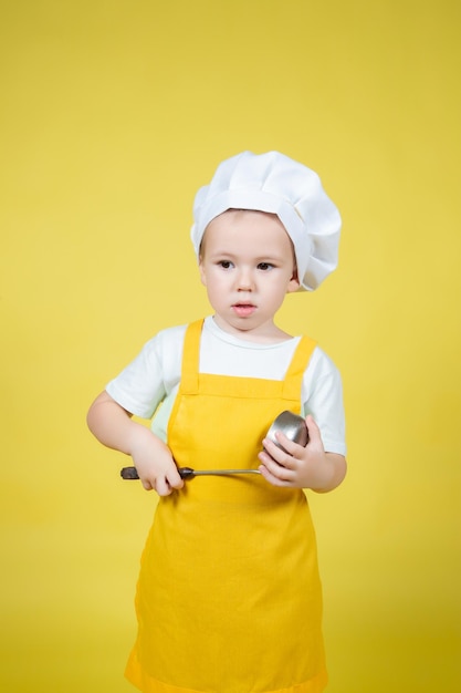 Kleine blanke jongen die chef-kok speelt, jongen in schort en koksmuts die emotioneel op gele achtergrond poseert