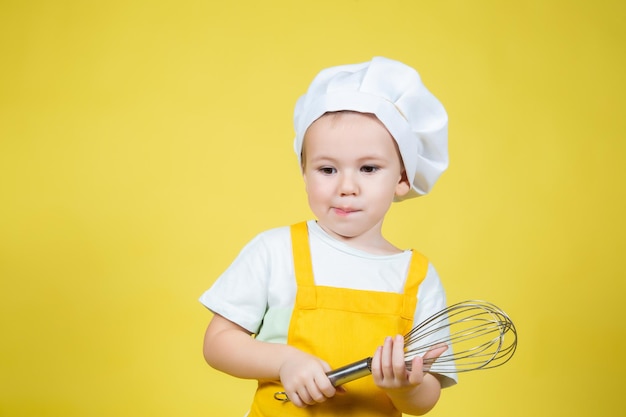 Kleine blanke jongen die chef-kok speelt, jongen in schort en koksmuts met een garde voor slagroom op gele achtergrond