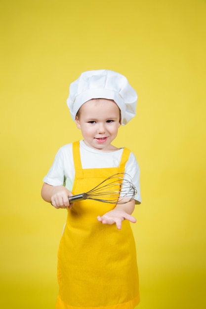 Kleine blanke jongen die chef-kok speelt, jongen in schort en koksmuts met een garde voor slagroom op gele achtergrond