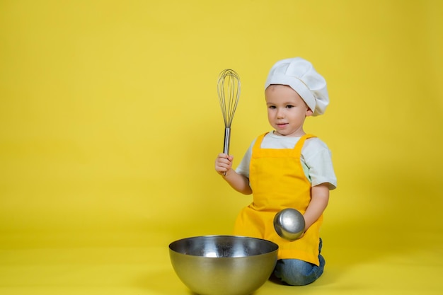 Kleine blanke jongen die chef-kok speelt, jongen in schort en koksmuts zittend op de vloer met een kom en zwaai op gele achtergrond