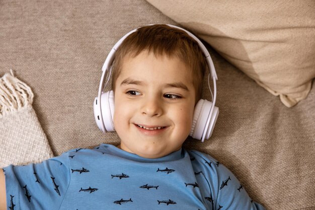 Kleine blanke jongen met blauw shirt en witte koptelefoon luistert naar muziek of audioboek op de bank