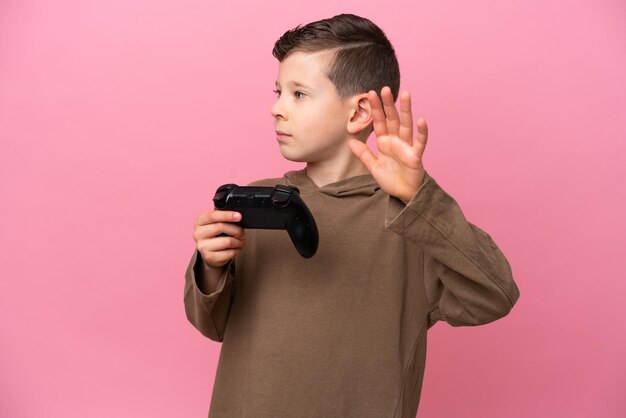 Foto kleine blanke jongen speelt met een videogamecontroller geïsoleerd op een roze achtergrond die een stopgebaar maakt en teleurgesteld is