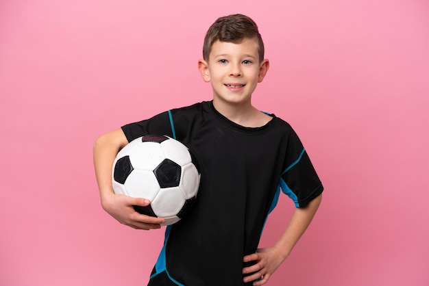 Foto kleine blanke voetballer jongen geïsoleerd op roze achtergrond poseren met armen op heup en glimlachend