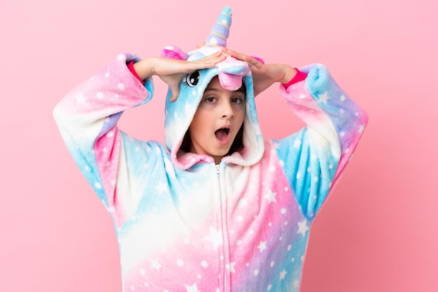 Kleine blanke vrouw draagt een eenhoornpyjama geïsoleerd op een roze achtergrond met verrassingsuitdrukking