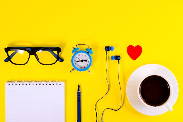 Kleine blauwe wekker, rood hart, oortelefoons, brillen en notitieboek, pen, op gele achtergrond.