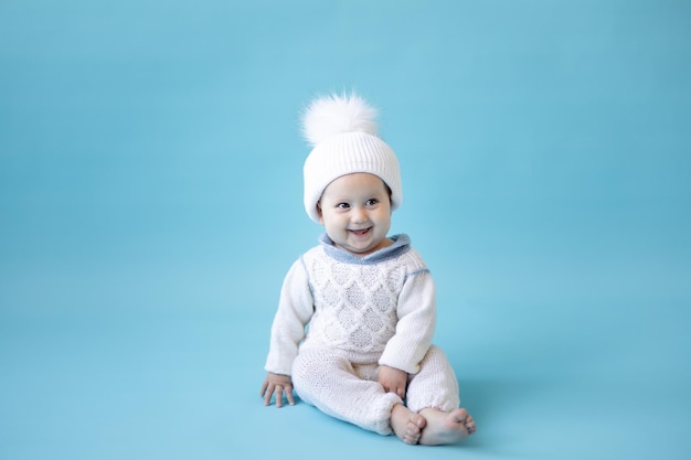 Kleine blonde babymeisje in een witte winter gebreide muts en gebreide trui op een blauwe achtergrond isoleer ruimte voor tekst baby peuter in winterkleren