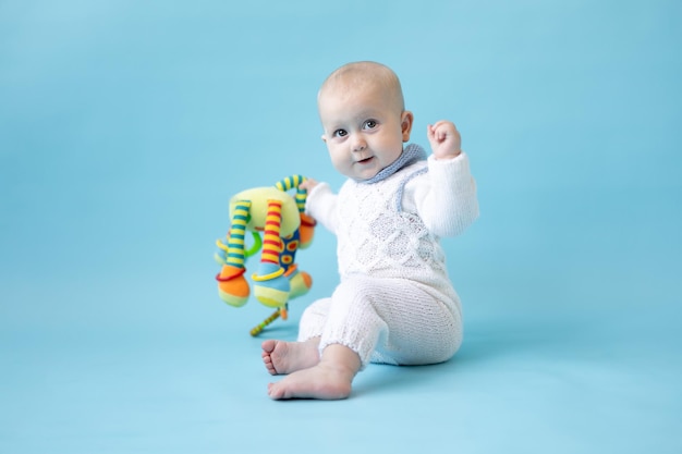 Kleine blonde babymeisje met speelgoed in haar handen in een gebreide trui op een blauwe achtergrond isoleren, ruimte voor tekst, baby peuter in winterkleren, winter