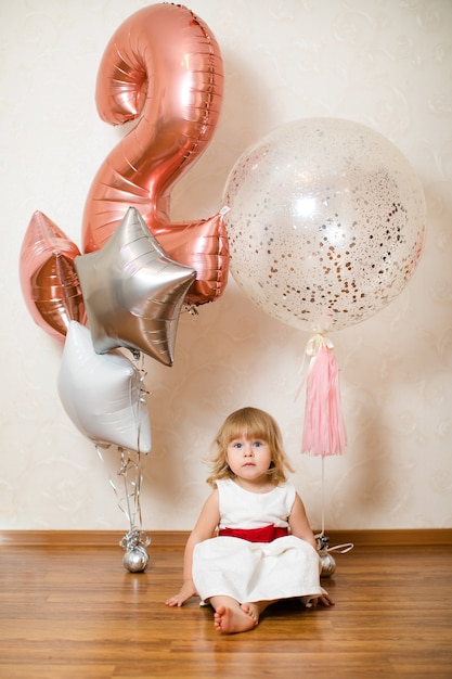 Kleine blonde babymeisje twee jaar oud met grote roze en witte ballonnen op haar verjaardagsfeestje.