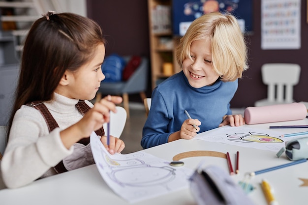 Kleine blonde jongen die naar een foto kijkt die is getekend en vastgehouden door zijn klasgenoot die hem hem laat zien tijdens de les van d