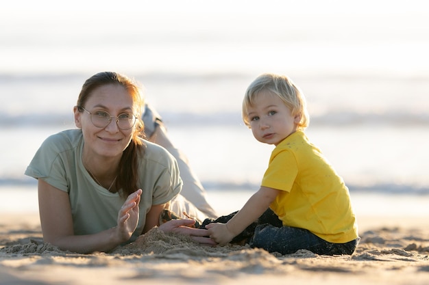 Kleine blonde jongen en zijn moeder spelen met het zand op het strand en kijken in de camera