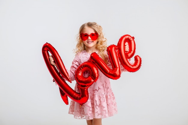 kleine blonde meisje in een roze jurk houdt in haar handen de inscriptie "liefde" van ballonnen op een witte muur