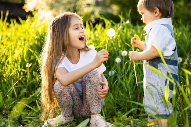 Kleine broer en zus in lichte zomerkleren. Leuk en grappig spelen met pluizige witte en gele paardebloemen tegen de achtergrond van hoog gras en weelderige groene bomen in de lentetuin.