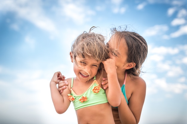 Kleine charmante zusjes fluisteren elkaar iets in het oor tijdens de zomervakantie op een warme zomerdag tegen een blauwe lucht