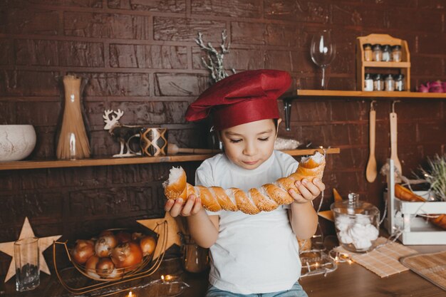 Foto kleine chef-kok. klein kind met een koksmuts met gebak, broodjes, brood en bagels. jongen die een ongezuurd broodje eet.