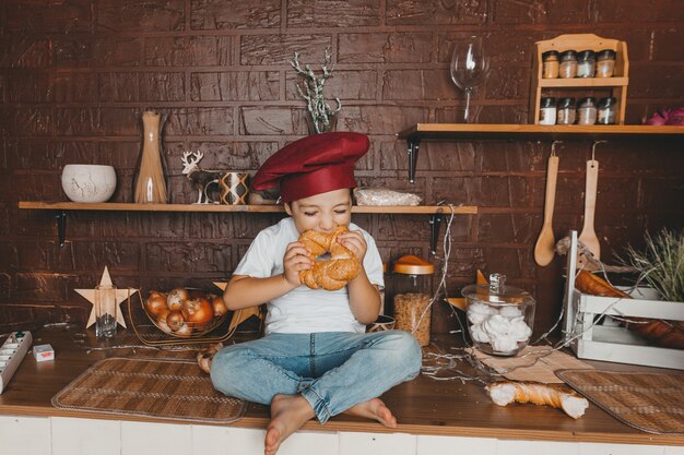 Kleine chef-kok. klein kind met een koksmuts met gebak, broodjes, brood en bagels. jongen die een ongezuurd broodje eet.