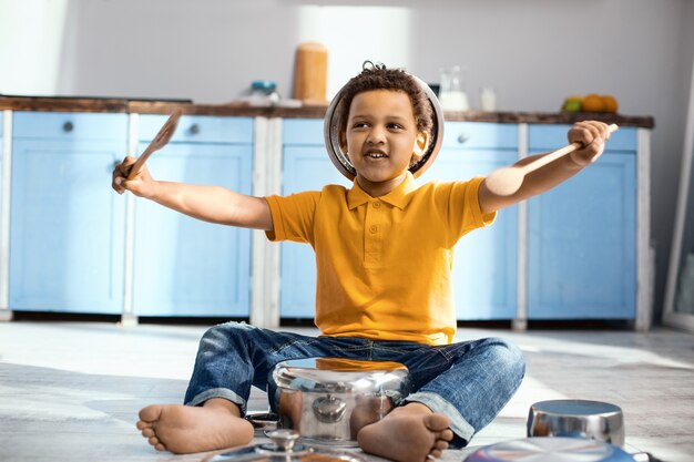 Kleine drummer. Vrolijke kleine drummer die op de keukenvloer zit en doet alsof hij een drummer is terwijl hij op een steelpan speelt