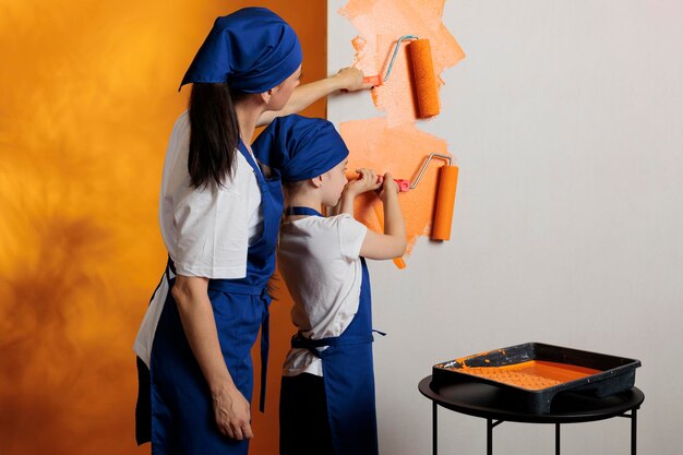 Kleine familie die muren schildert met oranje verf en rolborstel, die samen huishoudelijk werk doen. Moeder met jong kind met penseel en opknapgereedschap in appartement.