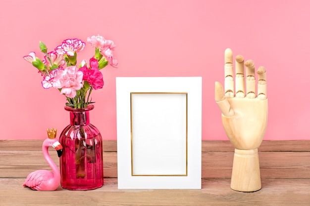 Kleine gekleurde roze anjers in vaas, witte fotolijst, figuur van flamingo, houten hand op houten tafel en roze muur