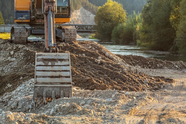 Kleine gele graafmachine op stapel stenen en stenen naast de rivier, detail op graafemmer op de grond. Bouw aan de rivier.