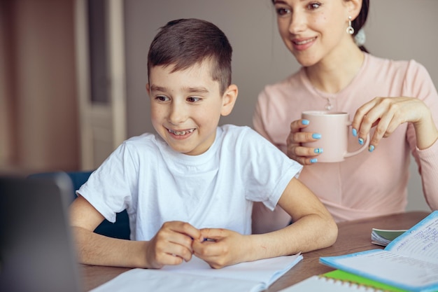 Kleine gelukkige jongen met bretels glimlachend en kijkend naar laptop moeder die koffie drinkt