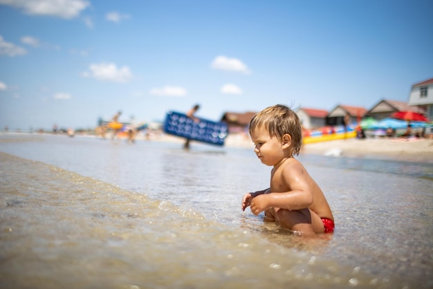 Kleine grappige grappige jongen, verzamelt schelpen en kiezelstenen in de kalme blauwe zee op een zandbodem onder de hete zomerzon op een heldere vakantie