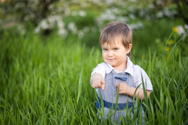 Kleine grappige jongen in een modieuze blauwe overall met expressieve blauwe ogen. Schattig lacht en eet vers groen gras in een grote bloeiende tuin in het hoge gras.