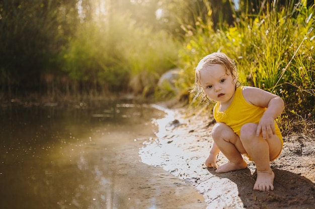 Kleine grappige schattige blonde meisjeskindpeuter in gele natte bodysuit die aan de waterkant van het meer speelt