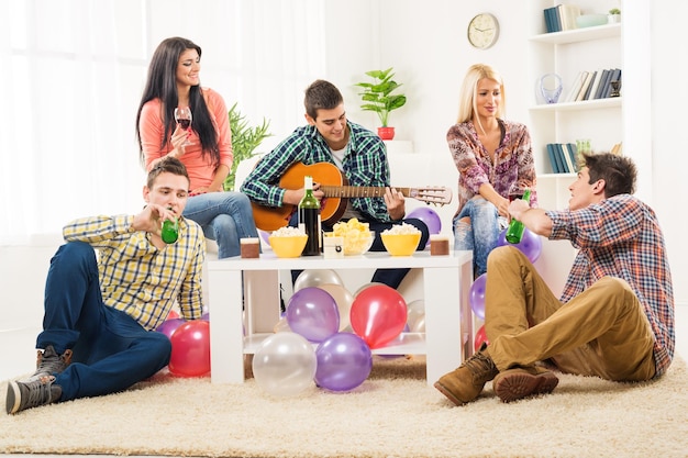 Kleine groep jongeren op het huisfeest, genieten van de klanken van de akoestische gitaar, wijn en bier drinken.