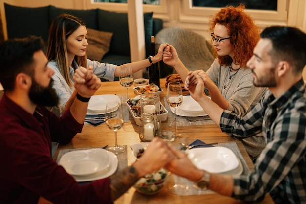 Kleine groep vrienden die elkaars hand vasthouden en bidden tijdens het eten aan de eettafel